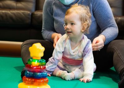 Photograph of baby girl sitting between her mother's legs, looking to the left and smiling.
