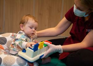 Photograph of baby looking towards, and playing with, a toy that a therapist is holding.