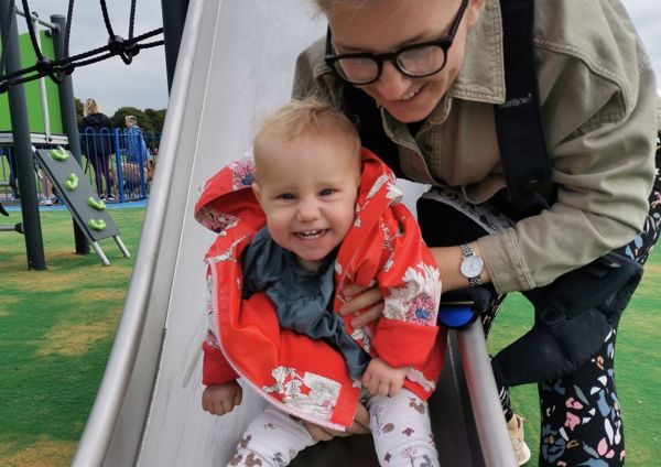 Photograph of a young girl at the bottom of a slide, smiling up at the camera whilst being supported by her mother.