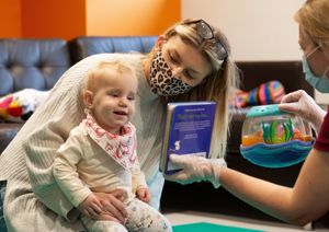 Photograph of baby girl sitting on her mother's lap, whilst looking up at a book that a therapist is holding towards her.