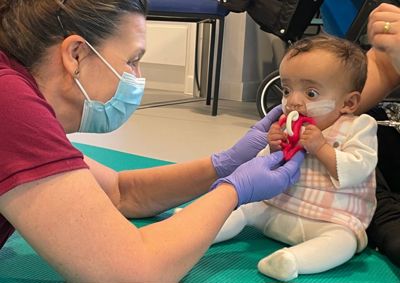 Photograph of baby girl putting a therapy toy into her mouth. Therapist on the left is passing the therapy toy to the baby.