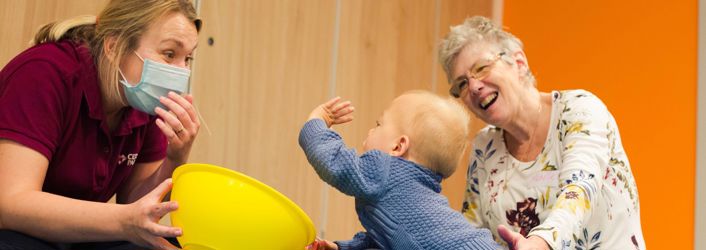 Photograph of baby boy reaching out towards a therapist. Supporting the baby on the right, is his grandmother, who is smiling at the baby.