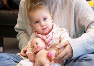 Photograph of baby girl holding a toy doll, looking up at the camera.