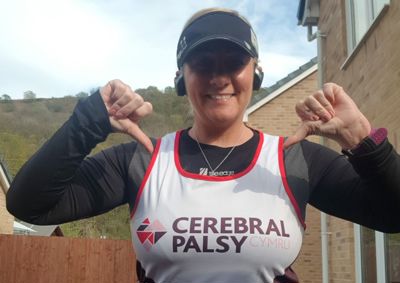Photograph of woman in Cerebral Palsy Cymru running vest, smiling at camera and pointing to her vest.