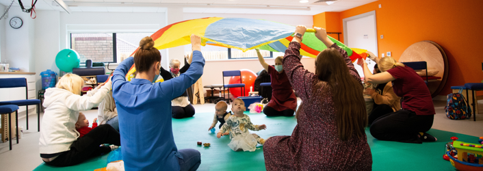 Photograph from Better Start playground, the adults are sitting in a circle holding up a parachute, whilst the babies sit underneath looking upwards towards the parachute.