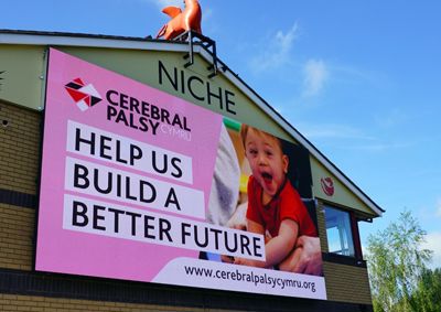 Photograph of a Cerebral Palsy Cymru branded billboard, that reads HELP US BUILD A BETTER FUTURE, on the building behind there is a Niche logo.