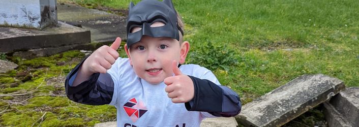 Photograph of young boy in batman mask and Cerebral Palsy Cymru branded t-shirt, looking at camera with his thumbs up.