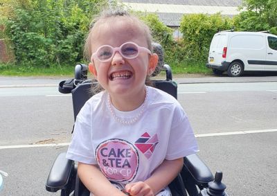 Photograph of a young girl in glasses and a cake & tea for cp t-shirt, She is sitting in her wheelchair and smiling at the camera.