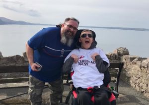 Photograph of man in a wheelchair (Rhys) alongside his support worker Andre. Rhys is wearing a Cerebral Palsy Cymru t-shirt and sunglasses. They are both smiling at the camera. 