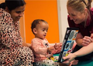 Photograph of a baby girl sitting in front of her mother, looking and reaching towards a mirror which a therapist is holding up.