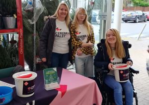 Photograph of two women standing, and one in a wheelchair, holding a bucket collection in a shop.