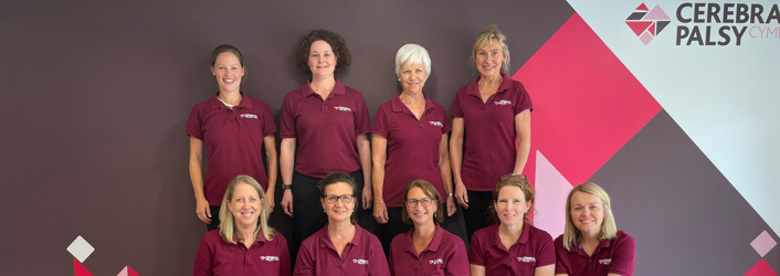 Photograph of therapy team (9 women) some seated, some standing, in front of Cerebral Palsy Cymru branded wall in our centre.