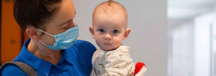 Photograph of woman in a mask holding a baby, who is looking directly at the camera.