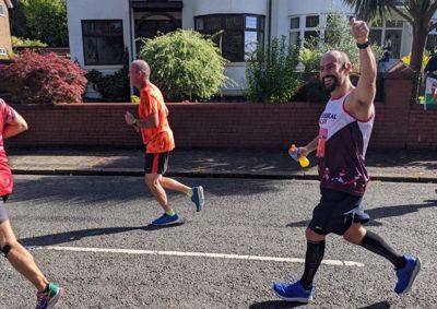 Photograph of Gareth, in his Cerebral Palsy Cymru running vest, running the Cardiff Half and waving at the camera.