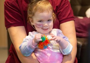 Photograph of baby girl holding a therapy toy, smiling at the camera.