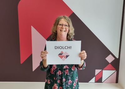 Photograph of Denise holding a Diolch sign with our tangram heart below it, she is standing in front of the branded wall in our centre.