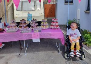 Photograph of young girl in a wheelchair smiling up at the camera, alongside her bake sale.