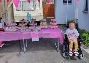 Photograph of young girl in a wheelchair smiling up at the camera, alongside her bake sale.