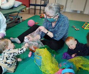 Photograph of woman (Glenys) holding out mesh fabric on the floor. One baby is reaching out towards the fabric Glenys is holding, another baby is looking at a different piece of fabric on the floor.