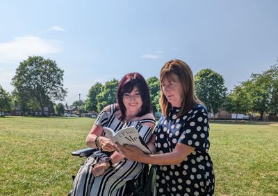 Photograph of Samantha Maxwell, alongside Lesley Griffiths with grass in the background. Both women are looking at the book 'CP Isn't me' which Lesley is holding out open in front of them.