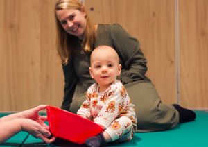 A baby sat on a therapy mat playing with a red plastic bowl 