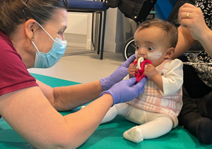 Photograph of baby girl putting a therapy toy into her mouth. Therapist on the left is passing the therapy toy to the baby.