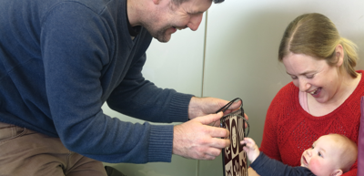 Baby in Mum's arms reaching out for therapy toy that dad is holding up.